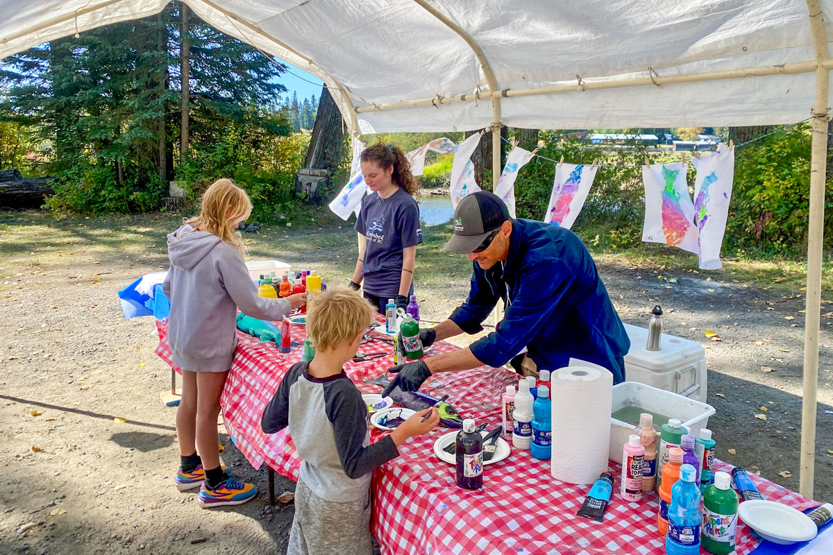 Two children are standing in front of a table covered with painting supplied and a red and white checkered tablecloth. Two Rivershed team members are standing behind the table and are helping the youth make salmon prints. Behind the two Rivershed members hang painted pictures of the Salmon.