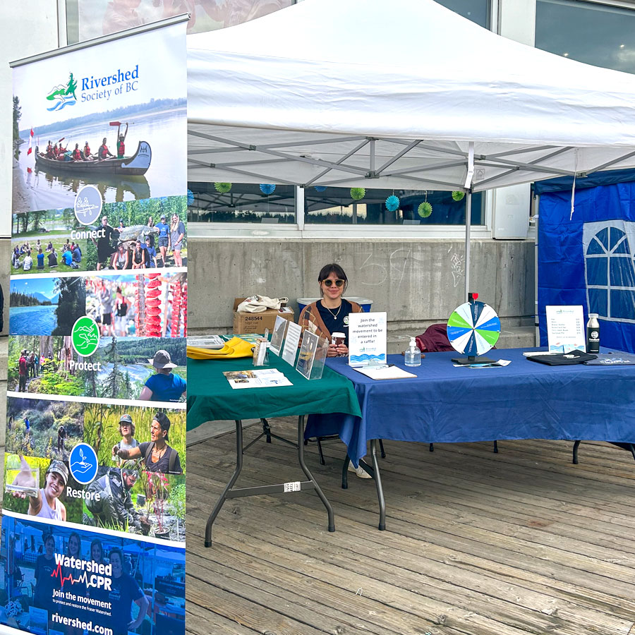 A female Rivershed team member is sitting behind two tables and smiling at the camera. There is a large banner in front of the table to promote Rivershed and shows photos from Rivershed’s programming. Promotional and informational material, merchandise, and an interactive spin-the-wheel game is on the table in front of the woman.