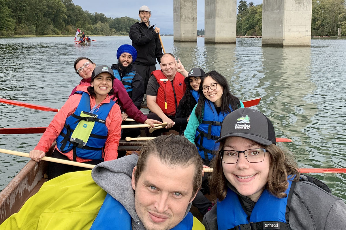 In the foreground of the photo are 8 paddlers and a skipper in the back half of a Voyageur canoe on the Lower Fraser River, smiling for the camera shortly after they pass under the Golden Ears Bridge between Langley and Pitt Meadows, B.C., on the shared territory of the Stó:lō, Kwantlen, Katzie, Semiahmoo, and Matsqui First Nations. The pilings of the bridge, a second Voyageur canoe and the tree line are shown in the background.