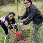625x625-CCSBCYouth12_7894Glover_October2023_LaraVolgyesi Two young woman are looking at the camera and smiling while they lower a sapling into a hole they dug in the ground.