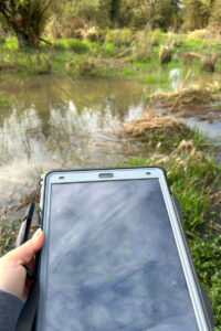 Hand holding a tablet in the foreground, the salmon river wetland surrounded by greenery and trees is visible in the background.