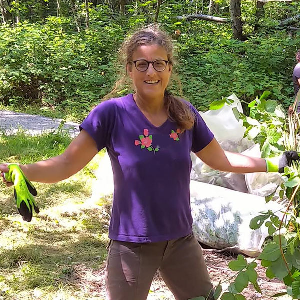 Woman in a purple shirt with long hair and glasses is standing with her arms out, gloves in one hand and invasive blackberries in the other hand.