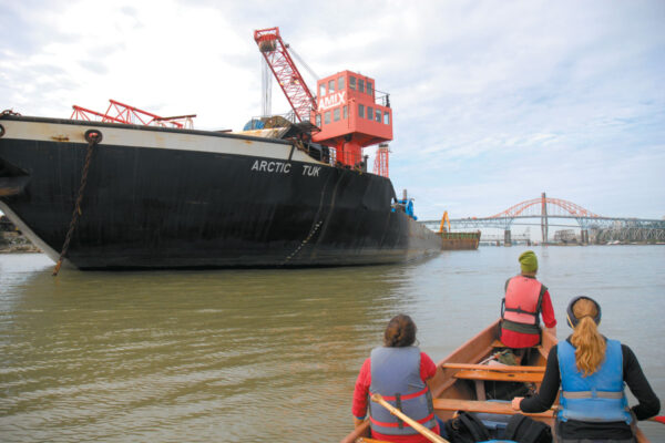 This photo is taken from the back of a Voyageur canoe. The backs of 3 people in the front of the canoe are visible, and they are beside a large cargo ship. The old Patullo Bridge is visible in the distance.