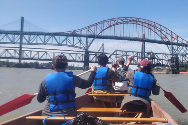Taken from the back of a large canoe, the backs of 4 people paddling and the canoe are in the foreground, with bridges in the background.